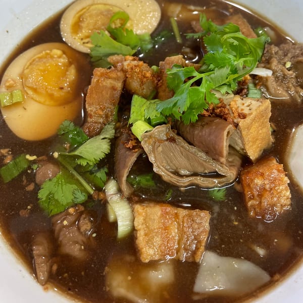 Close-up of Thai Kuay Jab Nam Sai soup with crispy pork, eggs, tofu, and cilantro.